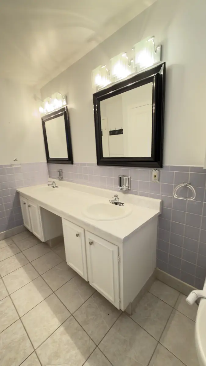 Dated lilac-tiled bathroom with a white builder vanity rebuilt with a walnut double vanity, gold fixtures and arched mirrors — before